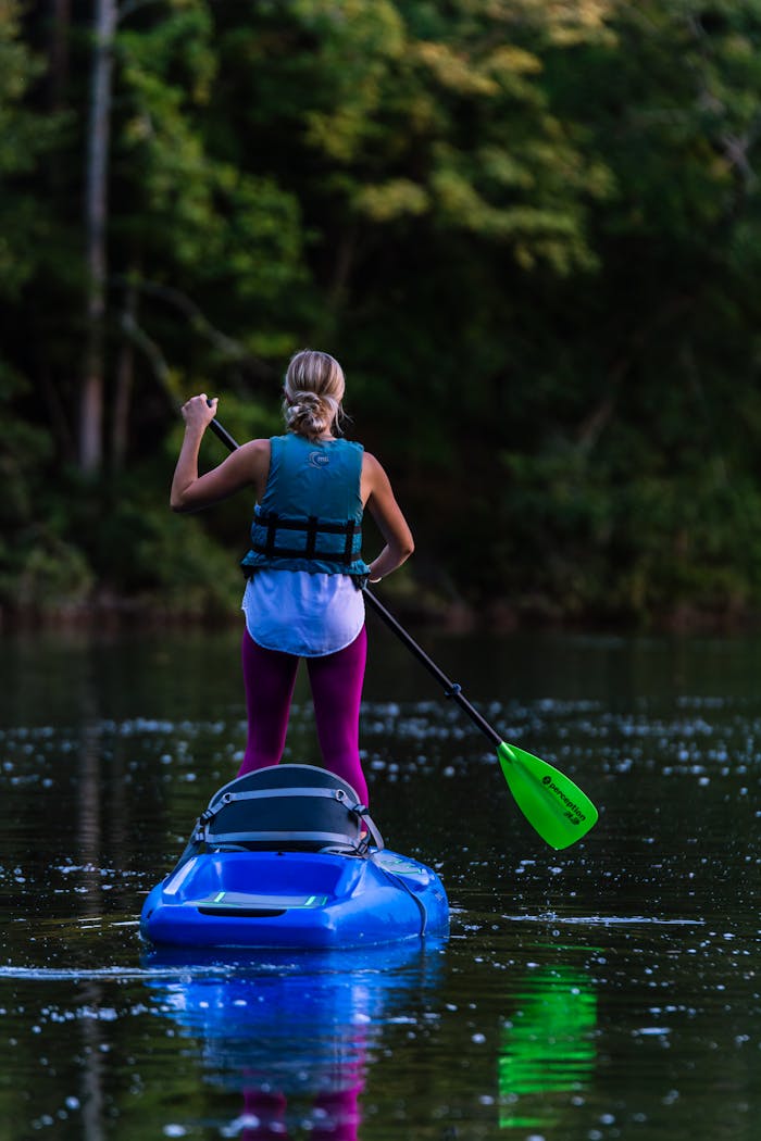 about-01 A woman enjoys stand-up kayaking on a calm lake surrounded by lush greenery, wearing a life vest.
