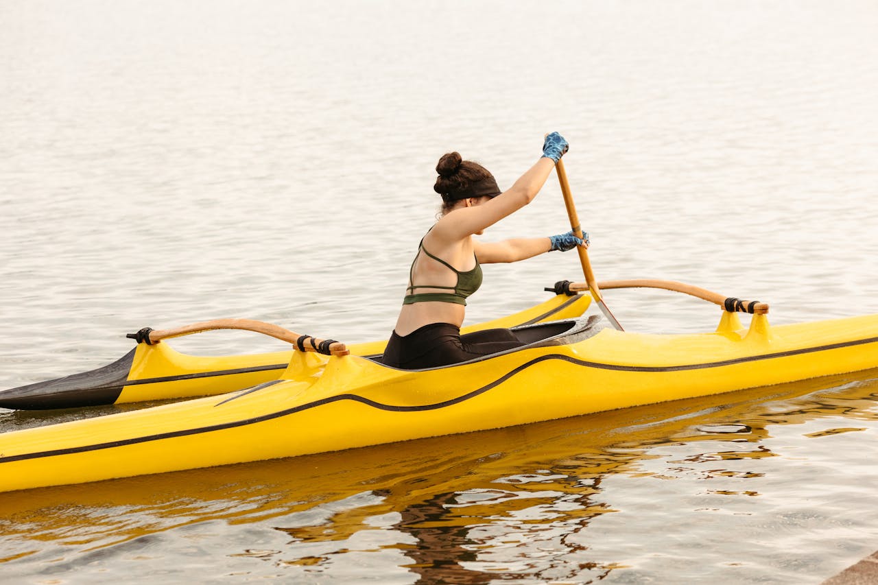 hero-img-01 A young woman paddles a yellow kayak on a peaceful lake during a bright summer day.