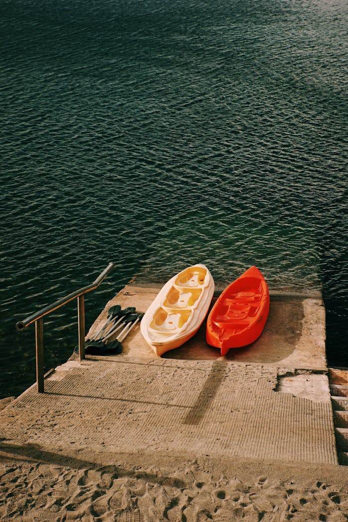 Two vibrant kayaks and oars by a slipway on a peaceful lake, ready for adventure.